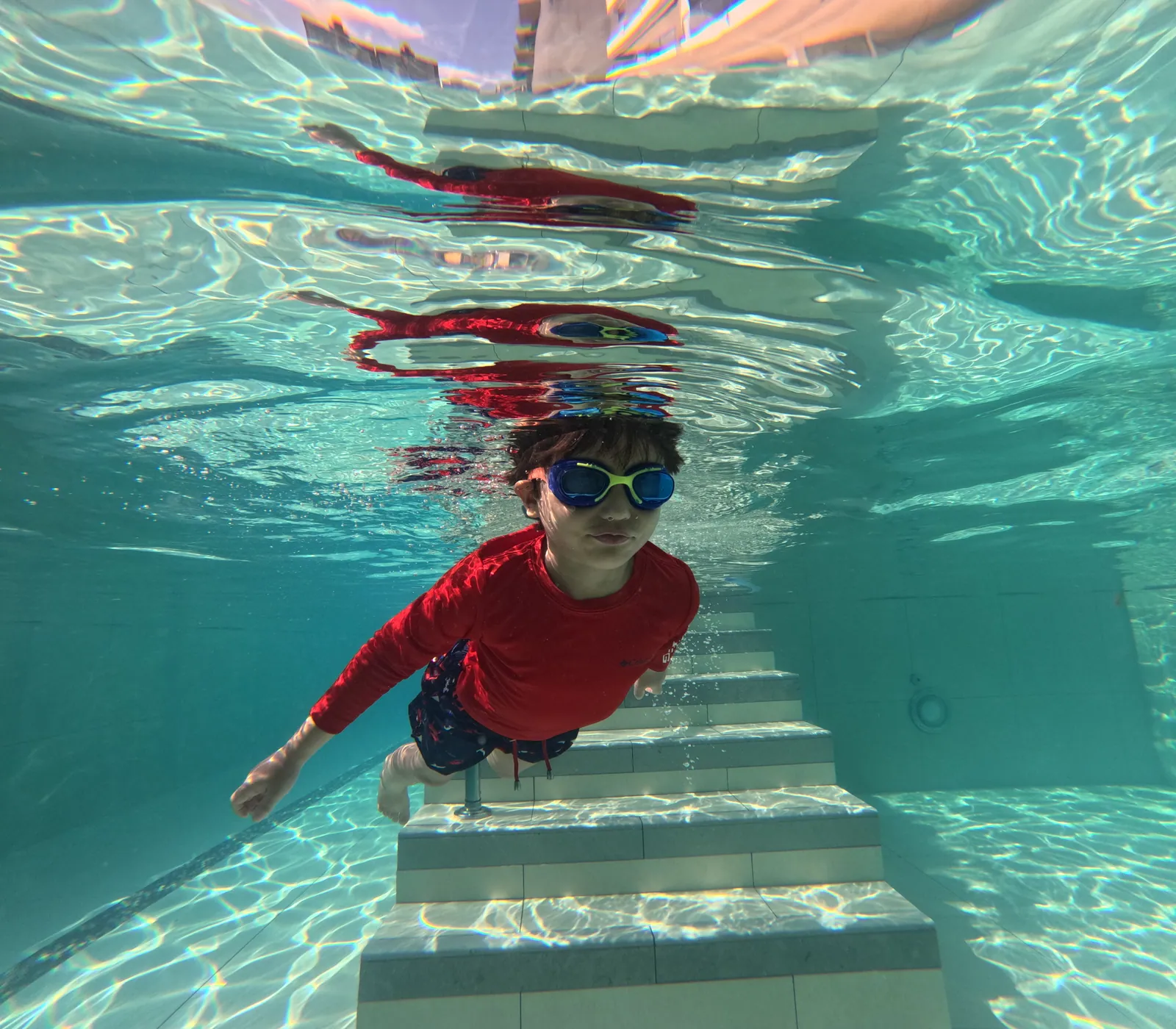 Niño con gafas nadando bajo el agua hacia las escaleras de la piscina