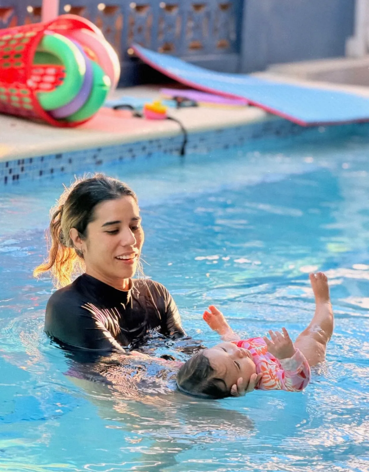 Instructora sosteniendo a un bebé que flota boca arriba junto a la piscina